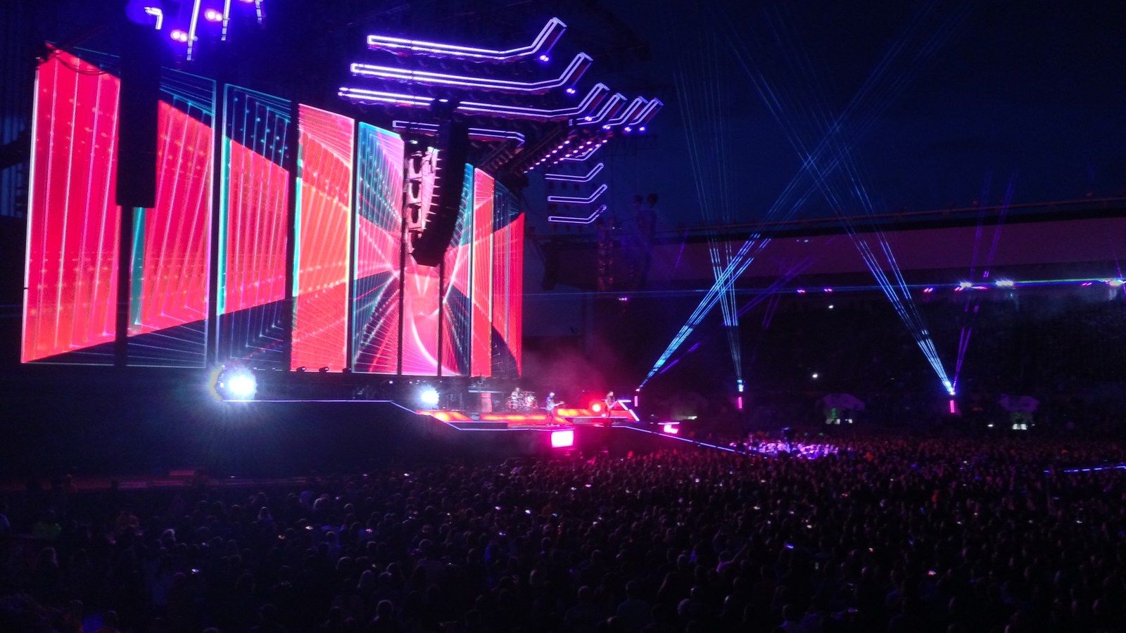 Stadium concert at night: a crowd in front of a stage of pink screens and blue light beams