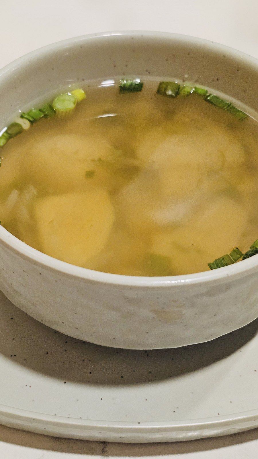 A bowl of clear broth with soft dumplings and pieces of potato, spring onions on top