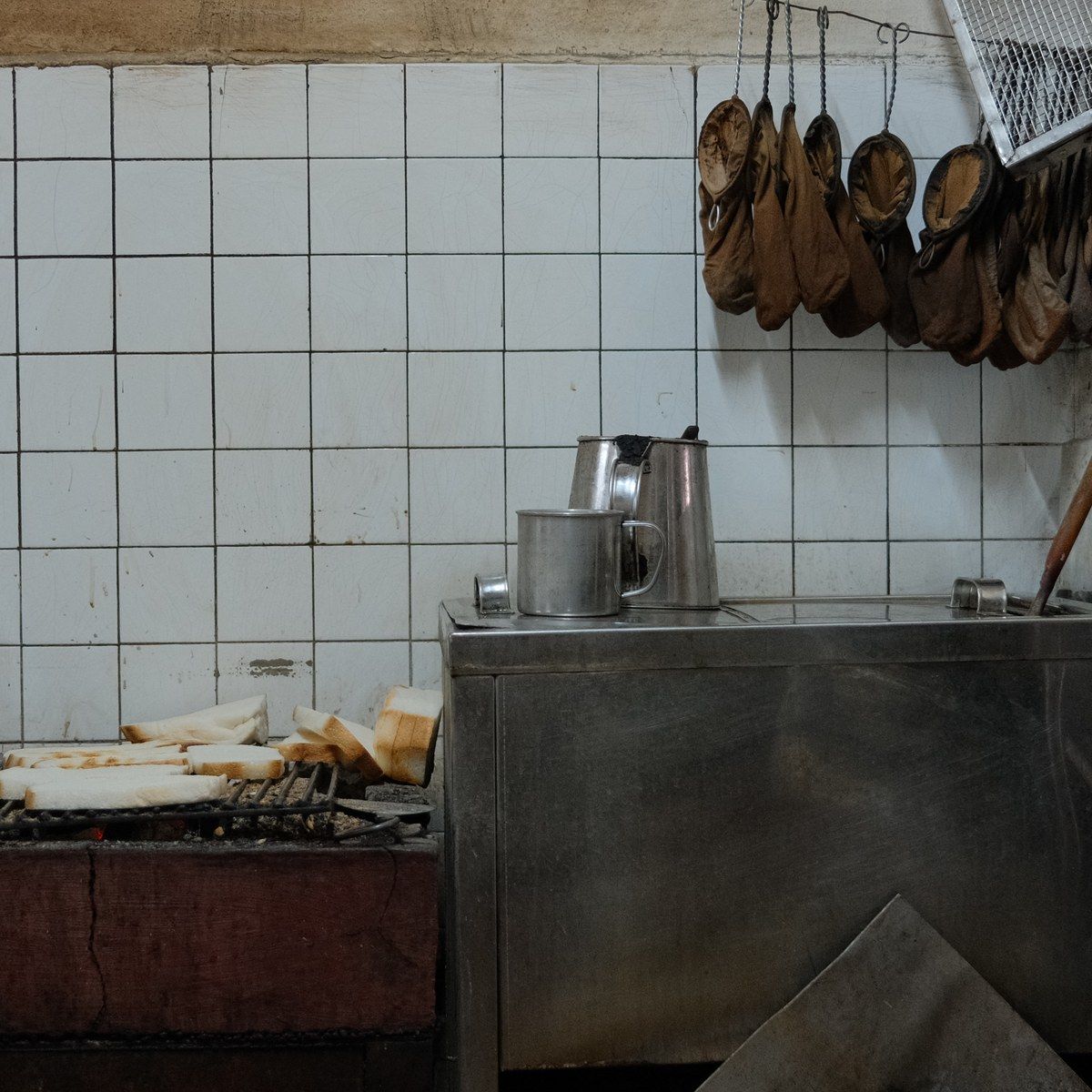 A small café kitchen with white tiles, bread toasting over charcoal and pans hanging on the wall