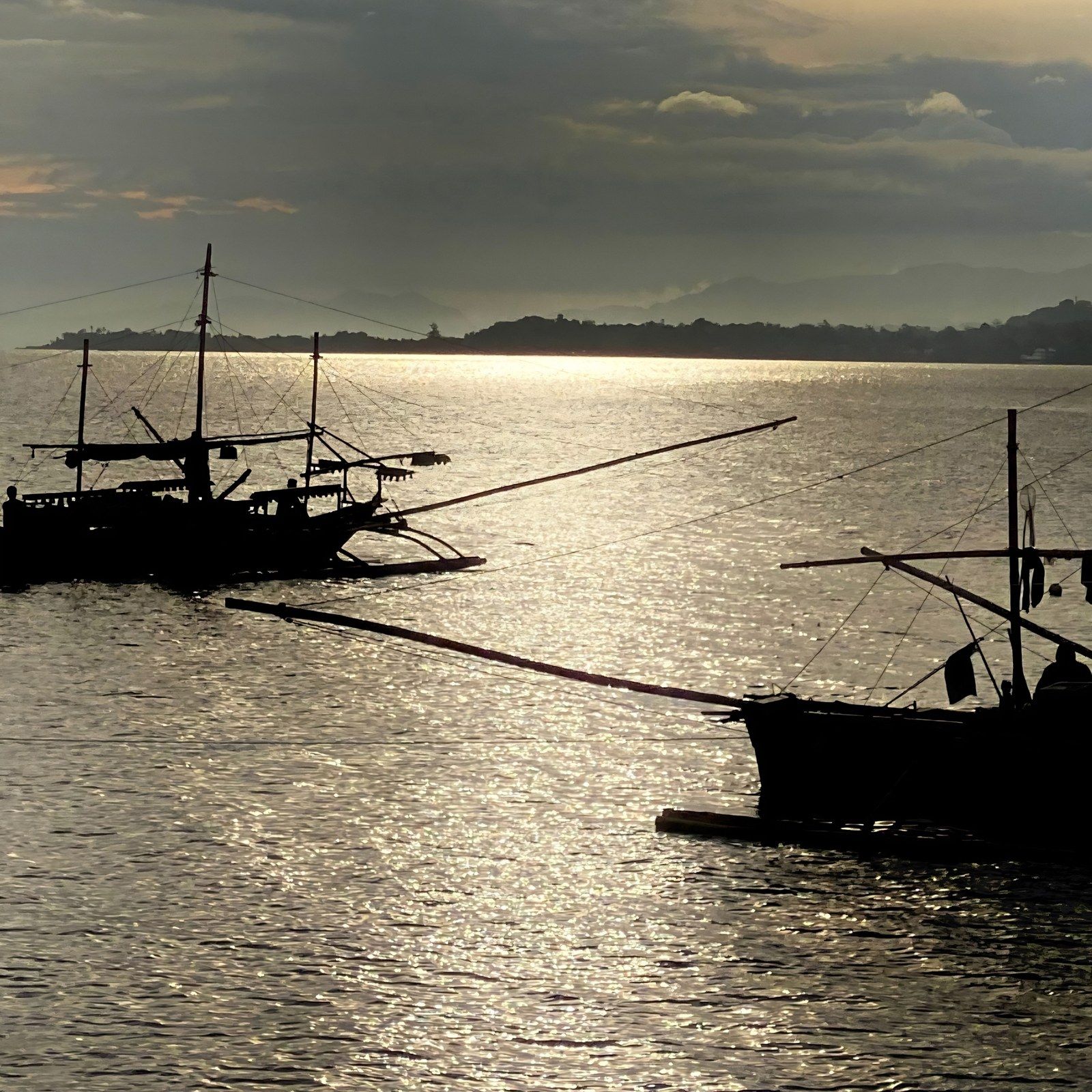 Two fishing boats crossing a calm sea against the light, their silhouettes on silver water