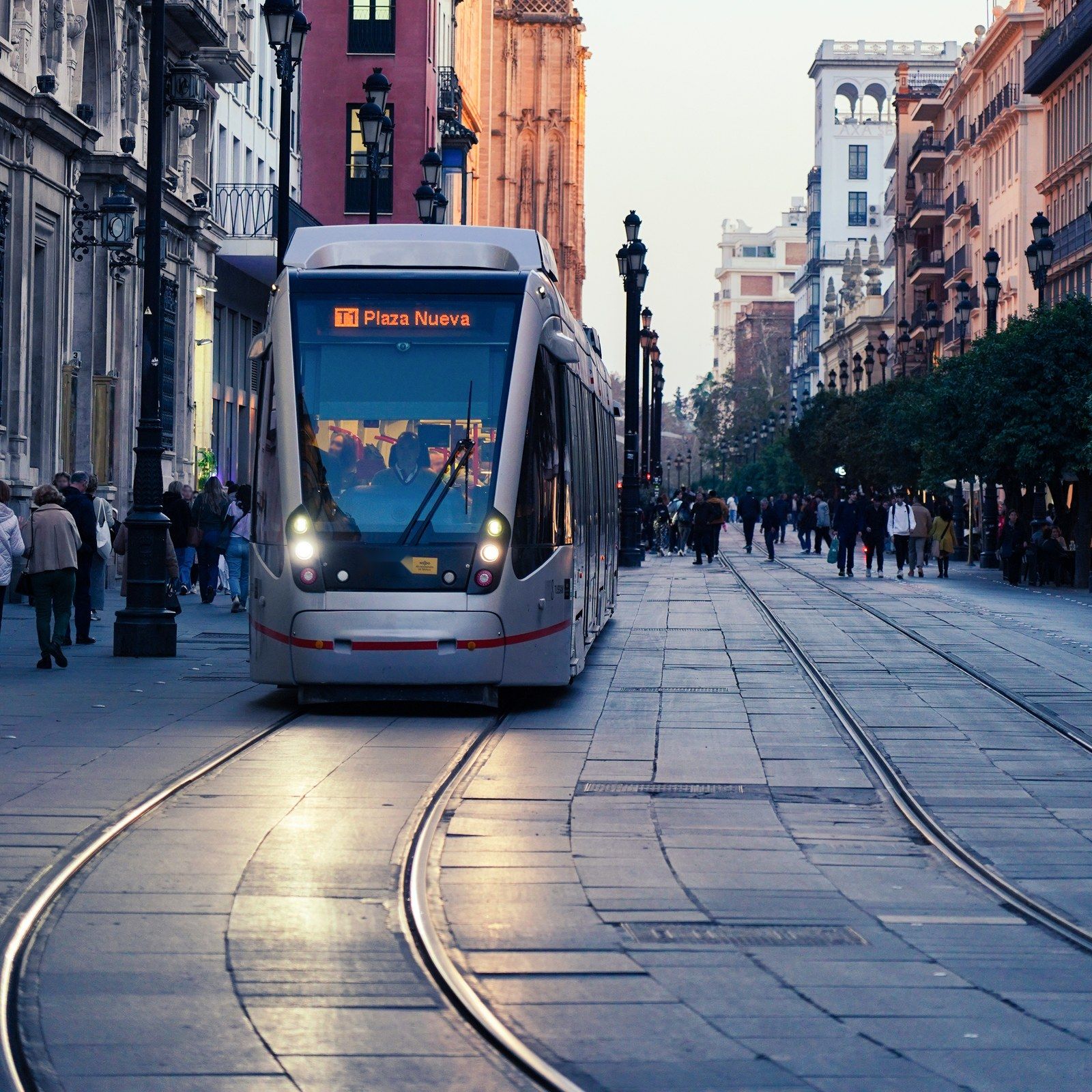 A modern tram coming down a wide pedestrian street between old buildings at dusk